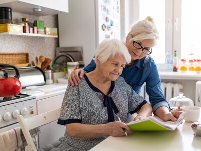 Rente für pflegende Angehörige: So sichern Sie Ihre Altersvorsorge | © Dr. Weigl GmbH & Co. KG Symbolbild zur Altersvorsorge – Miniatur-Senioren, Münzstapel, Haus und Uhr auf Kalender als Metapher für Rentenanspruch pflegender Angehöriger | © Dr. Weigl GmbH & Co. KG