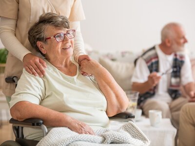 Tagespflege für Senioren – Betreuung und Entlastung für Angehörige | © Dr. Weigl GmbH & Co. KG Seniorin in Tagespflege-Einrichtung im Rollstuhl mit Pflegekraft und weiteren Senioren im Hintergrund | © Dr. Weigl GmbH & Co. KG